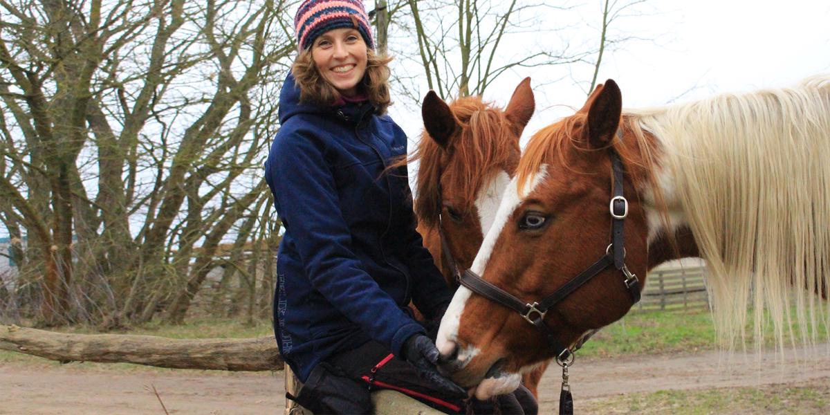 Dr. Anne Zinke, Landestierschutzbeauftragte des Landes Brandenburg Dr. Anne Zinke, Landestierschutzbeauftragte des Landes Brandenburg, lächelt in die Kamera, steht neben zwei Pferden und füttert dabei ein Pferd.