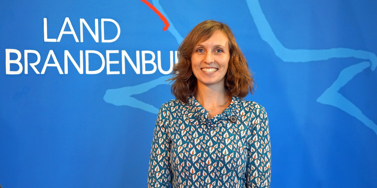 Tierschutzbeauftragte Dr. Anne Zinke Die Tierschutzbeauftragte Dr. Anne Zinke steht vor einer blauen Wand, auf der die Wort-Bild-Marke für das Land Brandenburg abgebildet ist.